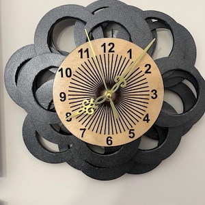 May include: A decorative wall clock with a light wood face and black outer design. The clock face features black radial lines and gold-colored hands. The outer design is made of overlapping black circles.