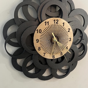 May include: A decorative wall clock with a unique design. The clock features a light wood-colored face with black numbers and gold-colored hands. The clock is surrounded by overlapping black circular shapes, creating a layered effect.