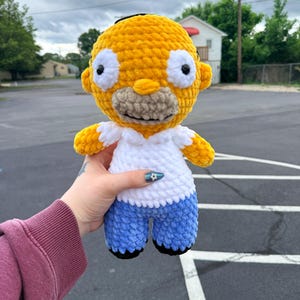 May include: A handmade crochet doll of a cartoon character with yellow skin, white eyes, and a tan mouth. The doll wears a white shirt and blue trousers. The doll is approximately 20 cm tall.