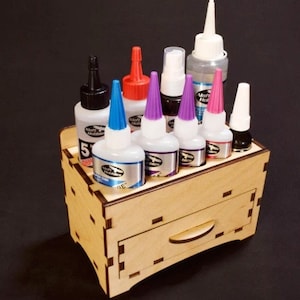 May include: A wooden organizer with a drawer for storing bottles of glue. The organizer has seven slots for bottles, and the bottles are various colors, including purple, pink, red, blue, and black.