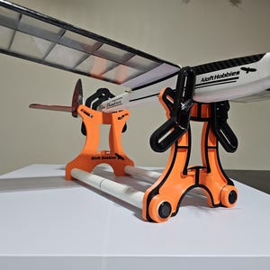 K&ouml;nnte beinhalten: Ein orangefarbener Flugzeugst&auml;nder aus Kunststoff mit zwei wei&szlig;en Rohren, der ein wei&szlig;-schwarzes Modellflugzeug mit dem Schriftzug "Aloft Hobbies" auf der Seite h&auml;lt.