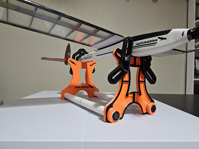 K&ouml;nnte beinhalten: Ein orangefarbener Flugzeugst&auml;nder aus Kunststoff mit zwei wei&szlig;en Rohren, der ein wei&szlig;-schwarzes Modellflugzeug mit dem Schriftzug "Aloft Hobbies" auf der Seite h&auml;lt.