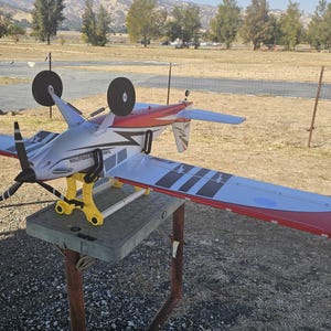 K&ouml;nnte beinhalten: Ein wei&szlig;es, rotes und schwarzes Modellflugzeug mit gelbem Fahrwerk steht auf einem Holzst&auml;nder. Das Flugzeug hat einen Propeller und befindet sich auf einem grasbewachsenen Feld.