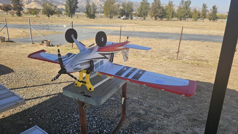 K&ouml;nnte beinhalten: Ein wei&szlig;es, rotes und schwarzes Modellflugzeug mit gelbem Fahrwerk steht auf einem Holzst&auml;nder. Das Flugzeug hat einen Propeller und befindet sich auf einem grasbewachsenen Feld.