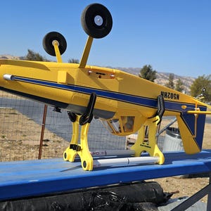 K&ouml;nnte beinhalten: Ein gelbes und blaues Modellflugzeug mit dem Text "NHZOSN" an der Seite steht auf einem blauen Tisch. Das Flugzeug steht auf einem gelben St&auml;nder mit zwei Beinen.