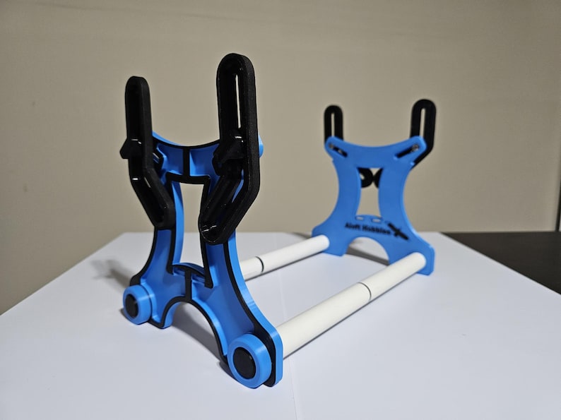 K&ouml;nnte beinhalten: Ein blauer und schwarzer 3D-gedruckter St&auml;nder mit zwei wei&szlig;en Stangen. Der St&auml;nder ist so konzipiert, dass er zwei Gegenst&auml;nde, wie z. B. Telefone oder Tablets, aufnehmen kann.