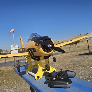 K&ouml;nnte beinhalten: Ein gelbes und schwarzes Modellflugzeug auf einem St&auml;nder mit einer schwarzen Fernbedienung. Das Flugzeug hat einen Propeller und steht auf einer blauen Oberfl&auml;che.