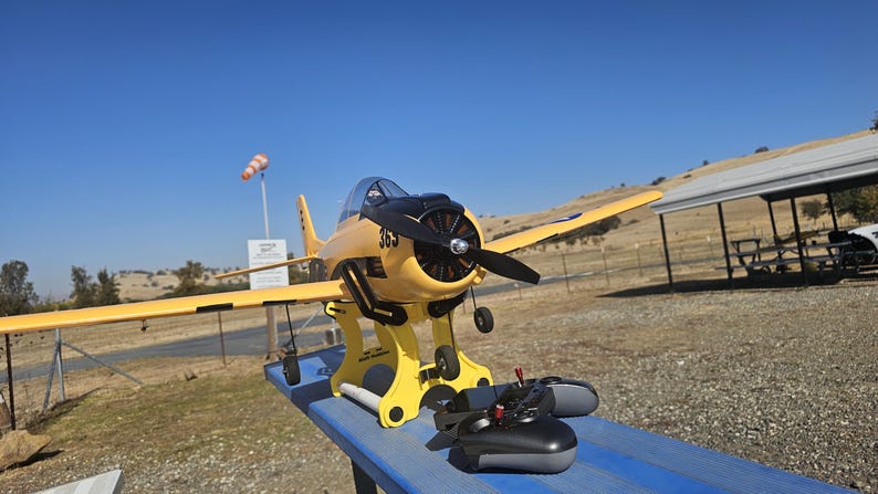 K&ouml;nnte beinhalten: Ein gelbes und schwarzes Modellflugzeug auf einem St&auml;nder mit einer schwarzen Fernbedienung. Das Flugzeug hat einen Propeller und steht auf einer blauen Oberfl&auml;che.