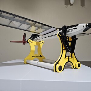 K&ouml;nnte beinhalten: Ein gelber Flugzeugst&auml;nder mit einem wei&szlig;en und schwarzen Flugzeugmodell darauf. Der St&auml;nder tr&auml;gt den Text "Aloft Hobbies". Das Flugzeugmodell tr&auml;gt den Text "Hobbie".