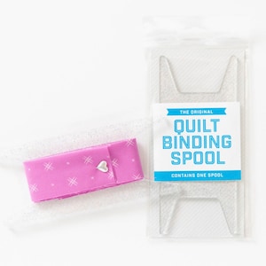 May include: A clear plastic spool for quilt binding, with a pink fabric binding already wound on the spool. The label reads "The Original Quilt Binding Spool. Contains One Spool."