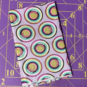 May include: A piece of fabric with a repeating pattern of colorful concentric circles on a light purple background.