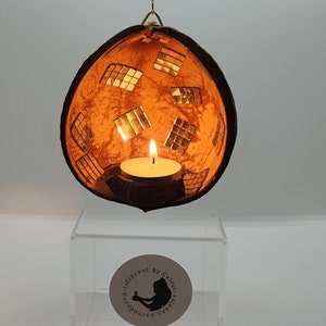 Peut inclure: Un bougeoir suspendu en forme de coque de noix de coco avec un motif miroir et une bougie chauffe-plat allumée à l'intérieur. Le bougeoir est suspendu à une chaîne dorée et repose sur une base en acrylique transparente. La base porte une étiquette qui indique "Life Scent by Celeste Handpoured Candles".