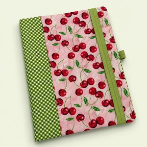 May include: A fabric-covered notebook featuring a pink background with a repeating cherry pattern and green leaves. The left side has a green and white gingham border, and a vertical green elastic band secures the cover on the right side.
