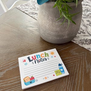 Puede incluir: Un bloc de notas cuadrado con el texto Lunch Notes en la parte superior. La p&aacute;gina presenta papel rayado con ilustraciones coloridas que incluyen un s&aacute;ndwich sonriente en un recipiente azul, una manzana roja, una botella de agua azul, una nota adhesiva amarilla y un sol.