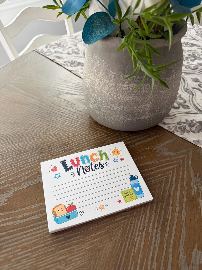 Puede incluir: Un bloc de notas cuadrado con el texto Lunch Notes en la parte superior. La p&aacute;gina presenta papel rayado con ilustraciones coloridas que incluyen un s&aacute;ndwich sonriente en un recipiente azul, una manzana roja, una botella de agua azul, una nota adhesiva amarilla y un sol.