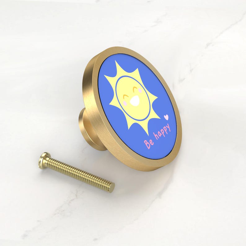 K&ouml;nnte beinhalten: Ein Messing-Schrankknopf mit einer blauen ovalen Fl&auml;che mit einem l&auml;chelnden gelben Sonnengrafik und den Worten "Be happy". Eine passende Messingschraube befindet sich neben dem Knopf. Dekorative Hardware.