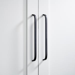 May include: Two black, curved cabinet handles fixed to a white cabinet door. The handles are long and slender, with a simple, modern design. The cabinet door is clean and minimalist, with a smooth, white surface. The handles are evenly spaced, adding a touch of elegance.