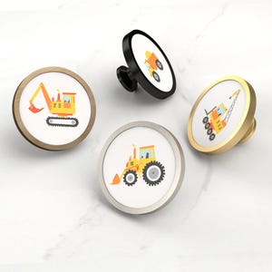 May include: Four round cabinet knobs with construction vehicle designs. The knobs feature illustrations of a yellow excavator, a yellow dump truck, a yellow tractor, and a yellow crane. The knobs have different metal finishes: gold, black, silver, and wood.