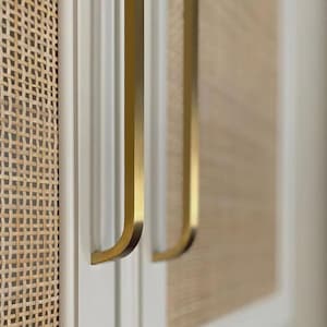 May include: Close-up of a cabinet door with two gold-colored, curved handles. The door is white with a woven rattan panel on the left and a similar panel on the right. The handles have a modern, minimalist design.