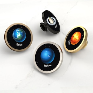 May include: Four cabinet knobs featuring planetary designs. The knobs include Earth, Neptune, Venus, and Saturn. Each knob has a black background with a planet image and a metallic border. The Earth knob has a wooden border, while the others have silver, gold, and black borders.