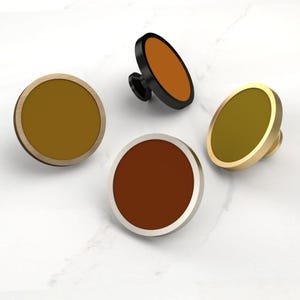 May include: Four circular cabinet knobs with flat faces and metallic rims. One knob has a black rim with an orange center, one has a brushed gold rim with a mustard center, one has a silver rim with a dark brown center, and one has a bronze rim.