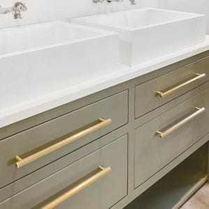 Puede incluir: Un mueble de baño moderno con dos lavabos rectangulares blancos y un mueble gris. El mueble tiene cuatro cajones con tiradores de barra dorados. La encimera es blanca y los grifos son plateados.