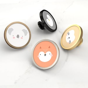 May include: Four decorative cabinet knobs with animal faces. The knobs feature a koala, a fox, a penguin, and a sloth. The knobs have a round shape with metal and painted details. The koala knob has a light brown frame, the fox knob has a silver frame.