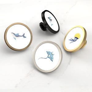 Peut inclure: Quatre boutons de meuble décoratifs avec des motifs de la vie marine. Les boutons présentent une baleine, un dauphin, une raie et une méduse. Chaque bouton a un fond blanc avec une illustration colorée et une finition métallique en or, noir, argent et bois.