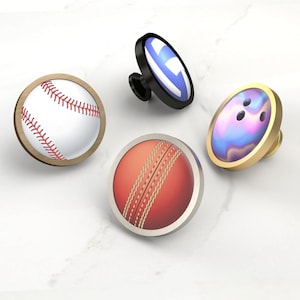 Op de afbeelding: Vier decoratieve kastknoppen met sportbalontwerpen. Inclusief een honkbal met rode stiksels, een cricketbal met stiksels, een bowlingbal met een gouden rand en een zwarte knop met een blauw-wit ontwerp.