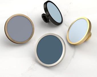 May include: Four circular cabinet knobs with flat faces and metallic rims. One knob has a brass rim with a slate blue center, one is black with a light blue center, one is silver with a dark blue center, and one is gold with a pale blue center.