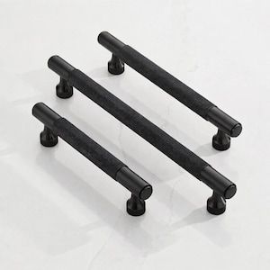 May include: Three black cabinet pulls with a textured, diamond-patterned grip. The pulls have a cylindrical bar and rounded ends, mounted on a curved base. The hardware is displayed on a white surface.