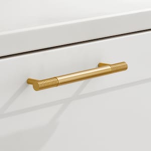 May include: A gold-colored cabinet pull with a cylindrical, ridged handle. The pull is mounted on a white cabinet door. The handle is approximately 6 inches long.