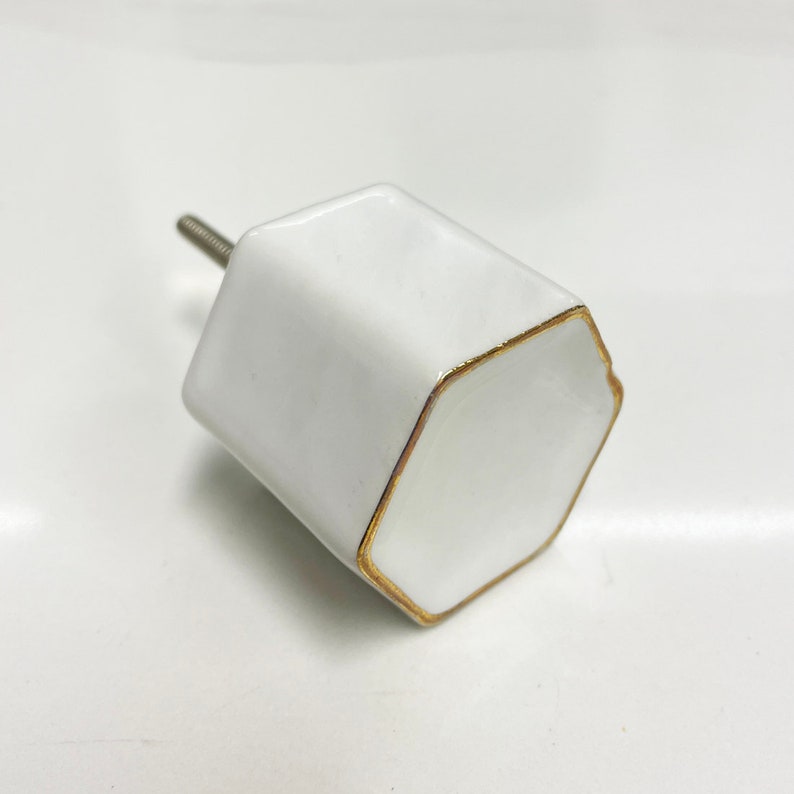May include: White ceramic cabinet knob with a gold trim around the edges. The knob has a hexagonal shape and a screw on the back.