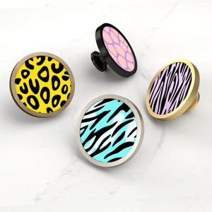 Könnte beinhalten: Vier dekorative Schrankknöpfe mit Tiermotiven. Die Knöpfe zeigen einen gelben Leopardenprint, ein rosa-lila Wabenmuster, einen blau-schwarzen Zebrastreifen und einen schwarz-weißen Zebrastreifen. Jeder Knopf hat eine andere Metallausführung.