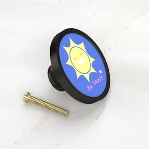 K&ouml;nnte beinhalten: Schwarzer Schrankknopf mit einem fr&ouml;hlichen Sonnendesign auf blauem Hintergrund. Die Sonne ist gelb mit einem l&auml;chelnden Gesicht und einem kleinen Herz. Die Worte "Be happy" sind in Pink geschrieben. Inklusive einer goldenen Schraube.