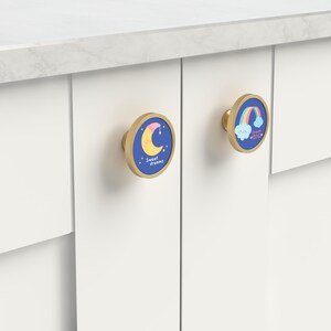 K&ouml;nnte beinhalten: Zwei Schrankkn&ouml;pfe mit goldfarbenem Finish und blauen Zentren. Einer zeigt einen Mond und die Worte "Sweet dreams". Der andere hat einen Regenbogen und die Worte "Dream BIG". Beide sind an einem wei&szlig;en Schrank montiert.