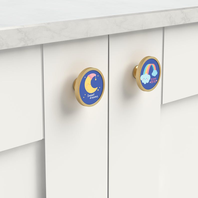 K&ouml;nnte beinhalten: Zwei Schrankkn&ouml;pfe mit goldfarbenem Finish und blauen Zentren. Einer zeigt einen Mond und die Worte "Sweet dreams". Der andere hat einen Regenbogen und die Worte "Dream BIG". Beide sind an einem wei&szlig;en Schrank montiert.