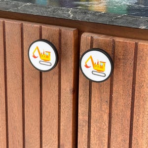 May include: Two circular cabinet knobs with a black rim and a white center featuring a yellow excavator graphic. The knobs are mounted on vertical wooden slats, providing a functional handle for opening and closing cabinet doors or drawers.