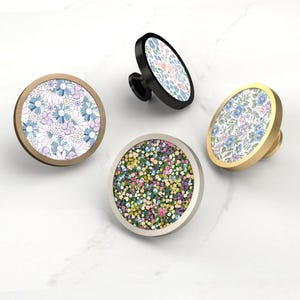 Op de afbeelding: Vier decoratieve kastknoppen met bloempatronen. Eén knop heeft een zwarte basis met een wit en blauw bloemmotief. Een andere heeft een gouden basis met een blauw en paars bloemmotief. De andere twee knoppen hebben een houten en zilveren basis met bloemmotieven.