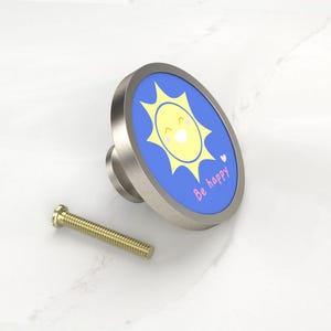 K&ouml;nnte beinhalten: Ein Schrankknopf aus geb&uuml;rstetem Metall mit einer blauen Oberfl&auml;che, die eine l&auml;chelnde gelbe Sonne und die Worte "Be happy" zeigt. Neben dem Knopf befindet sich eine goldfarbene Schraube.