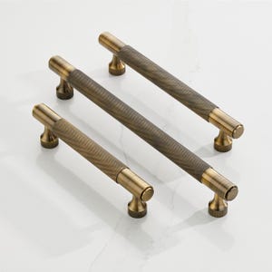 May include: Three antique brass cabinet pulls with a textured, diamond-patterned center and cylindrical ends. The pulls are arranged on a white surface, showcasing their warm, metallic finish. These decorative hardware pieces are ideal for kitchen or bathroom cabinets.