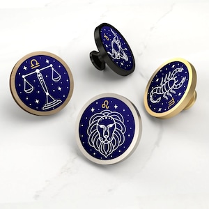 May include: Four cabinet knobs with zodiac designs. Each knob has a circular face with a dark blue background and a gold or silver rim. The zodiac symbols include Libra, Scorpio, and Leo, with white line art.