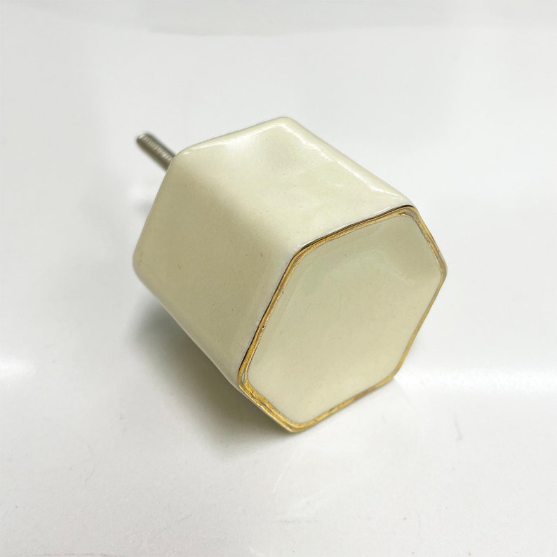 May include: A white ceramic hexagonal cabinet knob with a gold trim. The knob has a screw on the back for installation.