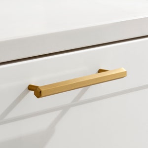 Può includere: Una maniglia per cassetto di armadio color oro con forma rettangolare e bordi leggermente arrotondati.