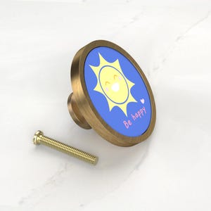 K&ouml;nnte beinhalten: Ein dekorativer Schrankknopf aus Messing mit einem ovalen blauen Gesicht, das eine l&auml;chelnde gelbe Sonne und die Worte "Be happy" in Rosa zeigt. Neben dem Knopf befindet sich eine passende Messingschraube. Der Knopf hat einen Durchmesser von ca. 3,8 cm.