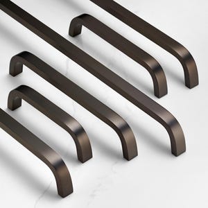 May include: Several bronze-coloured cabinet pulls arranged diagonally on a white surface. Each pull has a rectangular bar that curves into a U-shape. The pulls are made of metal and have a smooth, matte finish.