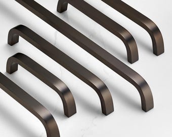 May include: A collection of bronze-colored cabinet pulls arranged diagonally on a white surface. Each pull has a rectangular bar that curves into a U-shape. The pulls are made of metal and have a smooth, matte finish.