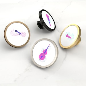 Pode incluir: Quatro puxadores decorativos para armários com desenhos de instrumentos musicais. Cada puxador apresenta uma face circular branca com um instrumento diferente em rosa e azul, incluindo uma guitarra, um microfone, um trompete e um violoncelo. Os puxadores têm acabamentos dourados, pretos, prateados e em tons de madeira.