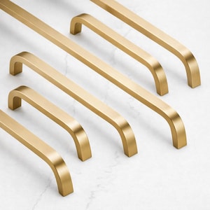 Könnte beinhalten: Eine Anordnung goldfarbener Schrankgriffe auf einer weißen Oberfläche. Die Griffe haben einen rechteckigen Stab mit gebogenen Enden, was eine moderne, minimalistische Ästhetik erzeugt. Geeignet für Küchen- oder Badezimmerschränke.