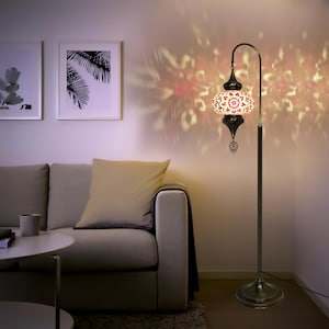 May include: A floor lamp with a white and pink mosaic glass shade. The shade has a geometric pattern and is accented with gold trim. The lamp has a curved arm and a gold base.